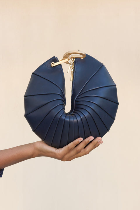 A navy blue leather clutch inspired by a sea shell design with a gold hardware interlocking-clasp closure, held up by a woman's hand. 