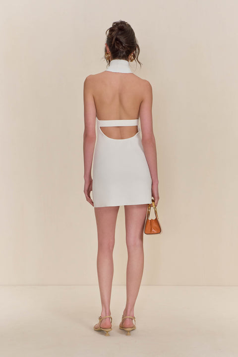 Back view of a woman wearing a white knit, sleeveless mini dress that features a high neck and low, open back with a strap across the back, paired with a tan handbag and sandals. 