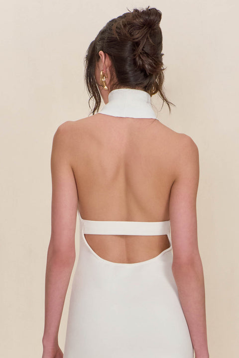 Detail shot of the open back detail on a white mini dress that features a high neckline and back strap detail, paired with chunky gold earrings. 