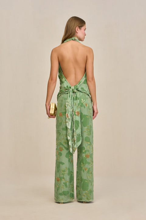 Back-facing woman wearing a green wide leg pant with velvet and silk burnout patterns and gold details, paired with a matching halter top with an open back.