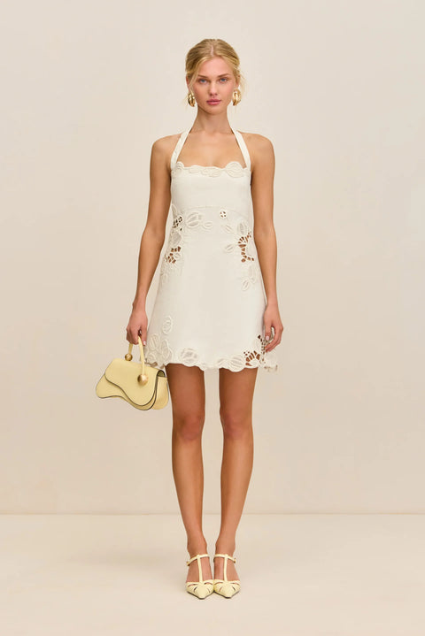 Woman wearing a white halter mini dress that features cutouts with floral lace overlays, paired with light yellow sandals and handbag. 