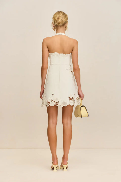 Back view of a woman wearing a white halter mini dress that features cutouts with floral lace overlays, paired with light yellow sandals and handbag. 