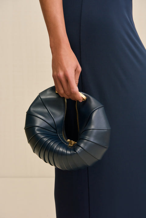 Woman holding a navy blue leather clutch inspired by the shape of a sea shell and finished with gold tone hardware and an interlocking clasp closure. 