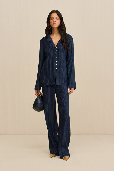 Woman wearing a navy plisse button down shirt that features pearlescent contrast buttons, paired with matching pants and a navy handbag. 