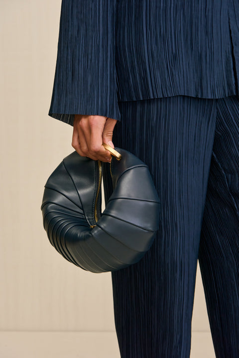 Woman holding a navy blue leather clutch inspired by the shape of a sea shell and finished with gold tone hardware and an interlocking clasp closure. 