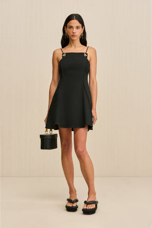 Woman wearing a black mini dress that features a square neck and grommet-style hardware on the spaghetti straps, paired with black sandals and a black purse. 