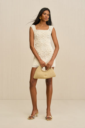 Woman wearing a sleeveless, white netted mini dress that features a corded floral overlay and square neck with a slip underneath for coverage, paired with heels and a woven handbag. 