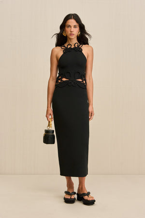 Woman wearing a black midi dress that features waist cutouts and floral piping overlays at the waist and neck. 