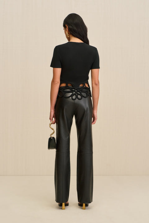 Back-facing woman wearing a black, knit t-shirt that features floral cutouts at the bottom hem, paired with black pants and black accessories. 