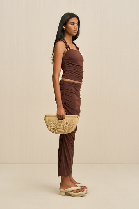 Side-facing woman wearing a brown tank top that features a knotted double strap detail and a matching brown maxi skirt, paired with tan sandals and handbag. 