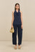 Front-facing woman wearing a navy blue sleeveless top that features a cowl halter neck, paired with blue pants, natural sandals, and a woven handbag. 