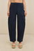 Close up of a woman wearing navy blue, utility style, barrel leg pants that features exaggerated pockets at the hips and on the calves, with gold-toned hardware, paired with tan sandals. 