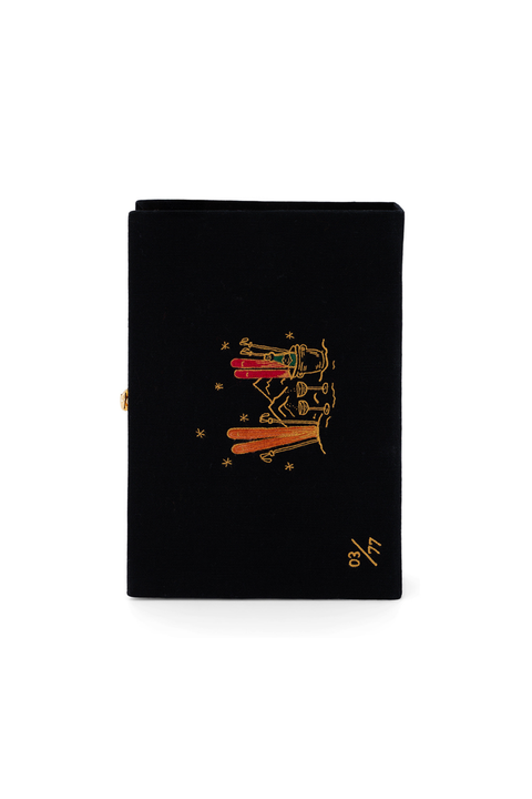 A black book clutch with a gold-toned metal clasp on the side and a gold-toned embroidered illustration of ski and champagne scene, set against a plain white background.