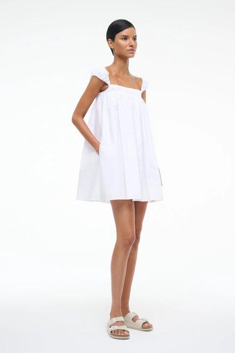 Side view of a woman wearing a white mini babydoll dress that features pockets and puffed cap sleeves. 