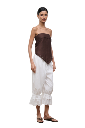 A model wearing a dark brown strapless top with silver studs and white ruffled cropped pants, standing against a plain white background.