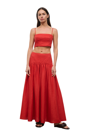 A woman modeling a bright red two-piece outfit consisting of a cropped spaghetti-strap top and a matching high-waisted maxi skirt, paired with black slide sandals, against a plain white background.