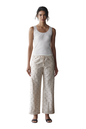 A woman modeling a two-piece loungewear set consisting of a white tank top and cream-colored wide-leg pants with a small black pattern, shot against a solid white background.
