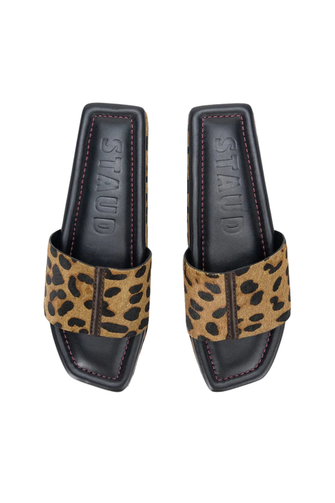 A pair of STAUD slide sandals featuring a tan and black leopard print strap on a black footbed with the brand name debossed, shown from a top-down view on a white background.