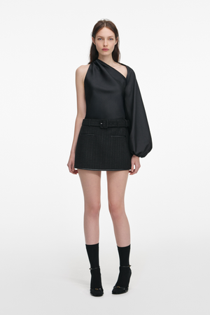 Woman wearing a black asymmetrical top and black mini skirt with belt with a white background