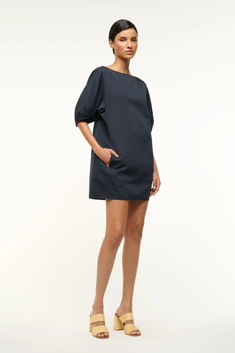 Side view of a woman wearing a navy, mini shift dress that features a boat neck and balloon half-sleeves, paired with tan heels. 