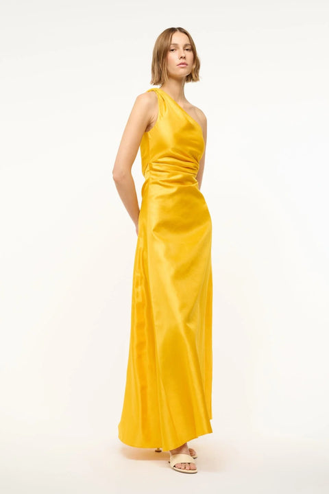 Side view of a woman wearing a yellow-gold silk maxi gown that features a one-shoulder detail with flattering draping at the bodice, paired with cream heels. 