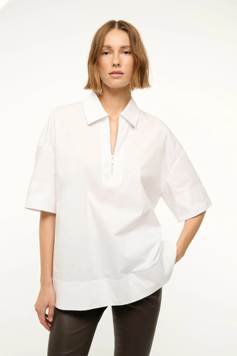 Close up of a woman wearing a white oversized short sleeve, collared tunic that features a zipper detail at the neck, paired with black capris.  