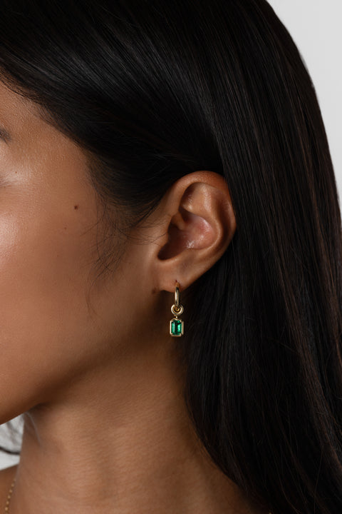 Woman wearing a small, yellow gold hoop earring that features three yellow gold annulets, one with an emerald cut emerald stone charm. 