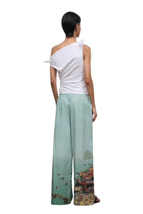 A model shown from the back wearing a white one-shoulder top and wide-leg trousers with a scenic coastal print in shades of green, blue, and orange.