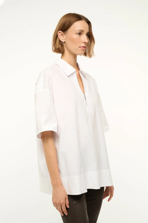 Side view of a woman wearing a white oversized short sleeve, collared tunic that features a zipper detail at the neck, paired with black capris.  