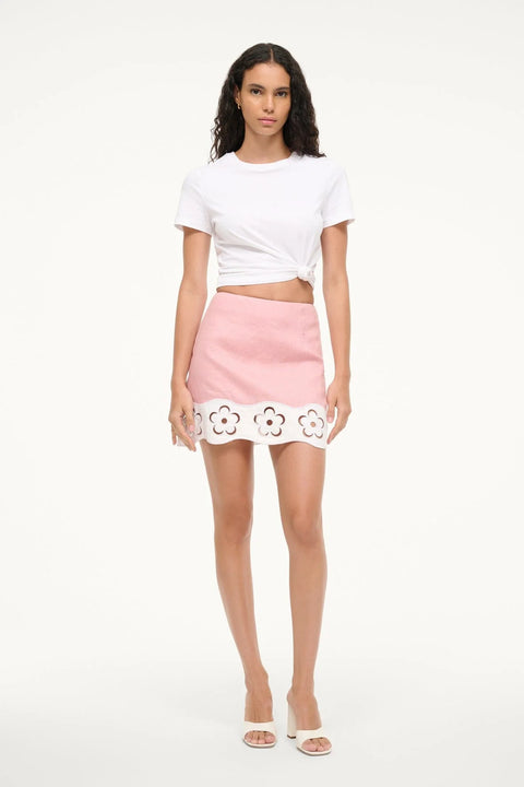 Woman wearing a pink linen mini skirt with a white contrast floral cutout hem, paired with a white top. 