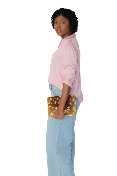 A woman in a light pink button-up shirt and light blue jeans holds a rectangular travel pouch featuring a brown and cream antilope pattern, shown against a plain white background.