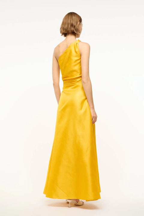 Back view of a woman wearing a yellow-gold silk maxi gown that features a one-shoulder detail with flattering draping at the bodice, paired with cream heels. 