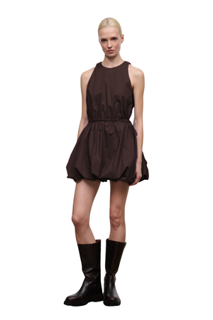 A model wearing a dark brown sleeveless mini dress with a gathered waist and puffed skirt, paired with black knee-high boots, set against a plain white background.