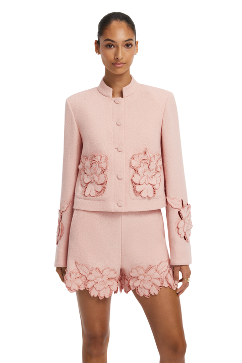 A model wearing a light pink two-piece suit featuring a cropped jacket with large 3D floral appliques on the chest and sleeves, paired with matching shorts that have a floral lace hem.