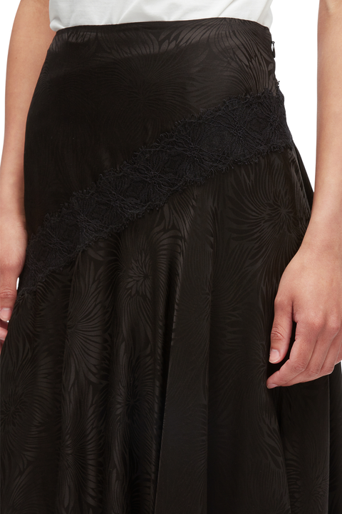 Close-up of a black skirt with a subtle tonal floral jacquard pattern and a wide black lace diagonal band across the front, shown on a model.