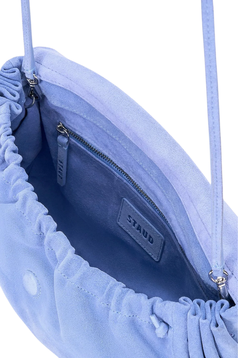 Close-up of the interior of a light blue suede shoulder bag with a drawstring closure and a zippered pocket. The brand name 'STAUD' is embossed on a leather patch inside.
