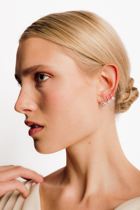 A model wearing a round diamond stud earring set in a white gold bezel suspended from a yellow gold stick in three different sizes. 