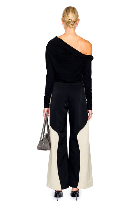 Woman facing away wearing a one-shoulder black top and high-waisted pants, holding a gray handbag.