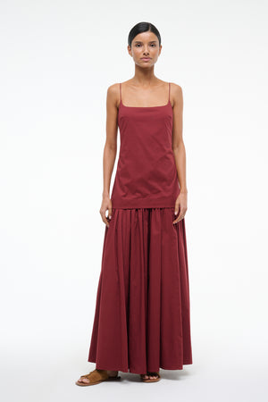 Woman wearing a deep red maxi dress that features a column bodice layered over a full skirt, paired with brown sandals. 