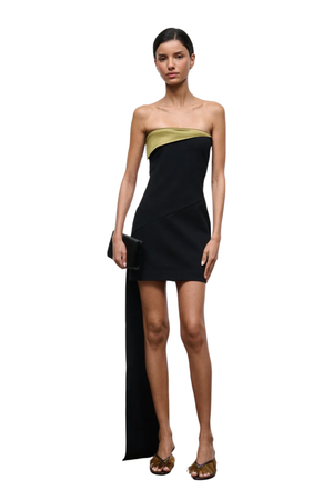 A model wearing a strapless black dress with a green band at the bust and a long draped side panel, paired with gold flat sandals and holding a black clutch, set against a plain white background.