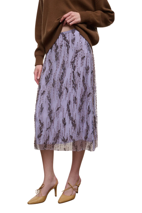A model wearing a lavender embellished midi skirt with a dark brown abstract vertical pattern, styled with a brown sweater and tan pointed-toe heels against a white background.