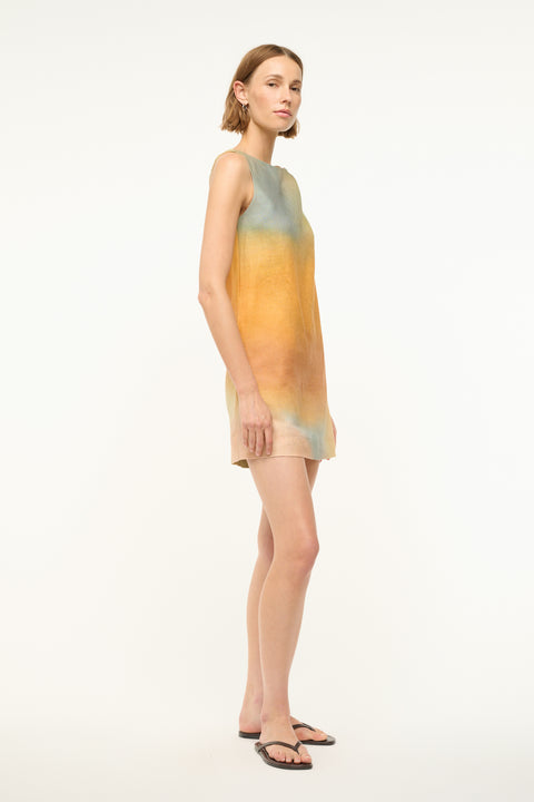 Side view of a woman wearing a sleeveless, pastel multicolor, mini shift dress that features a twist detail on one shoulder, worn with black sandals. 