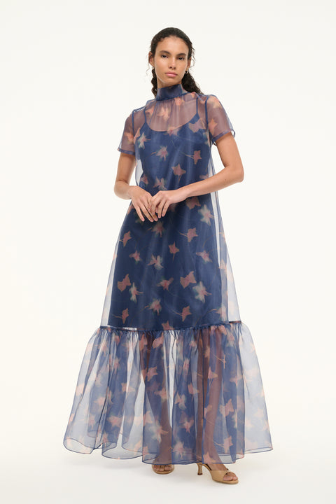 Woman wearing an organza gown in a navy floral print that features a mock neck, tiered ruffle hem, and removable slip, paired with tan heels. 