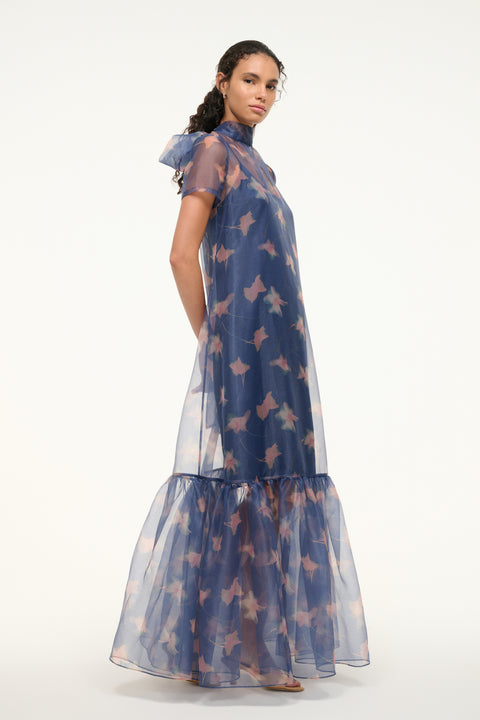 Side view of a woman wearing an organza gown in a navy floral print that features a tiered ruffle hem, removable slip, and mock neck with a bow at the back of the neck. 
