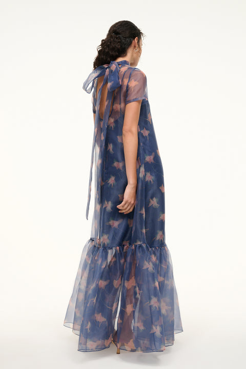 Back view of a Side view of a woman wearing an organza gown in a navy floral print that features a tiered ruffle hem, removable slip, and mock neck with a bow at the back of the neck. 