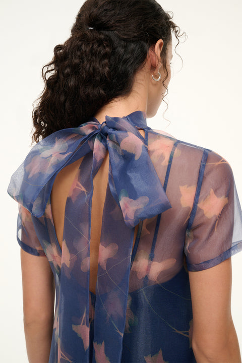 Detail shot of a woman wearing a short sleep organza gown in a navy floral print that features an open back with a mock neck that ties into a dramatic bow at the back of the neck.