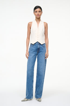 Woman wearing a cream button down vest that features a V neckline, paired with blue jeans. 