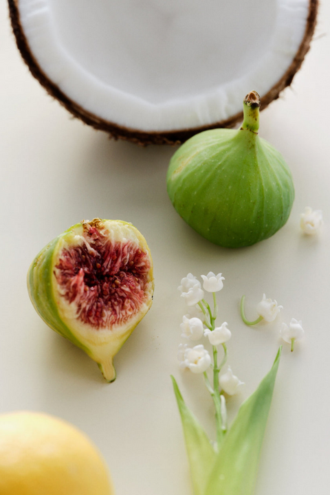 A styled flat-lay featuring a whole green fig, a halved fig showing pinkish-red seeds, a cut coconut half, a yellow lemon slice, and a lily of the valley flower stem on a light cream background.