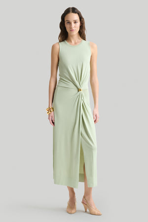 Woman wears light green maxi dress with gathering at waist with gold hardware and front slit.