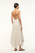 Back view of a woman wearing a v-neck ivory with brown stripes maxi dress that features a side pleat and ruching at the hip and and open back. 